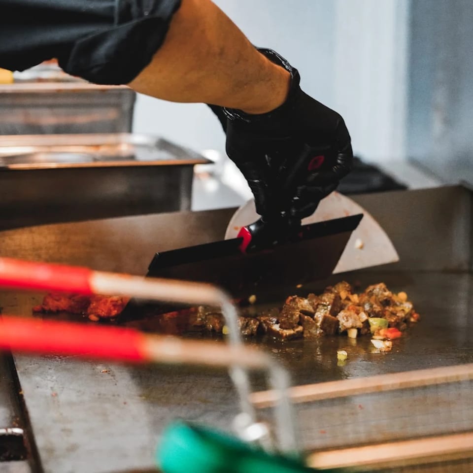 A chef in black gloves uses a spatula to cook diced meat and vegetables on a griddle.