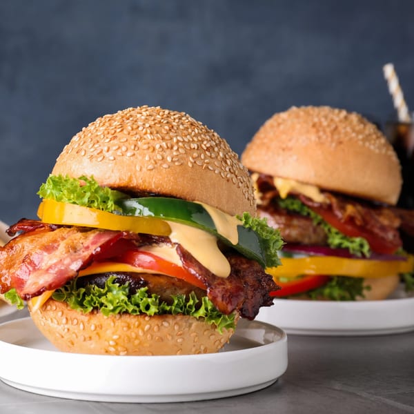 Two gourmet burgers with bacon, cheese, lettuce, tomato, and bell peppers on sesame buns.