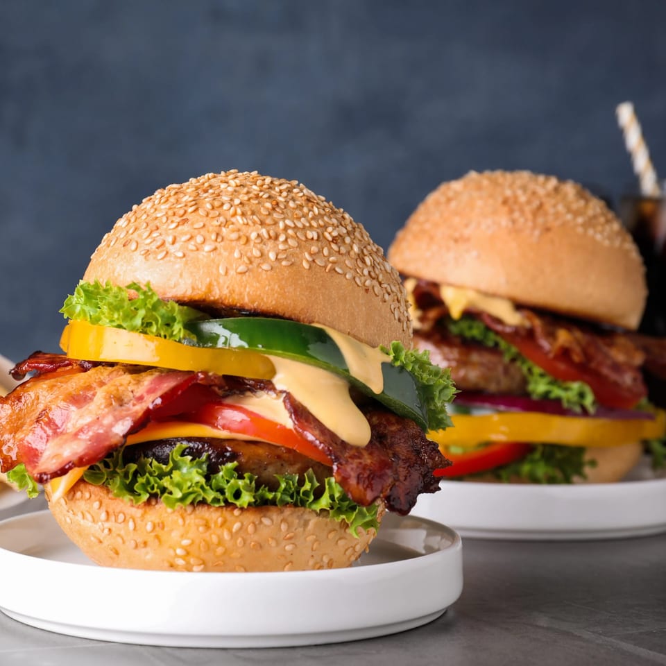 Two gourmet burgers with bacon, cheese, lettuce, tomato, and bell peppers on sesame buns.