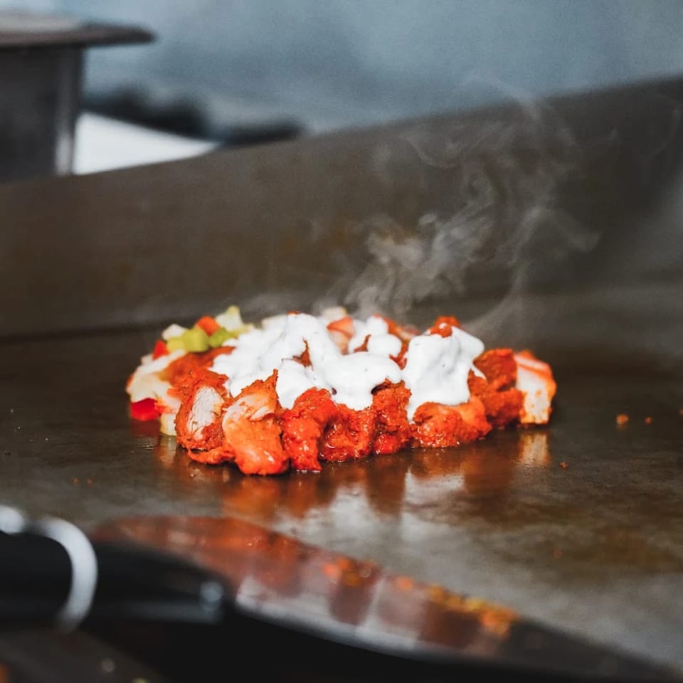 Sizzling chicken tikka with white sauce and vegetables cooking on a flat top grill.