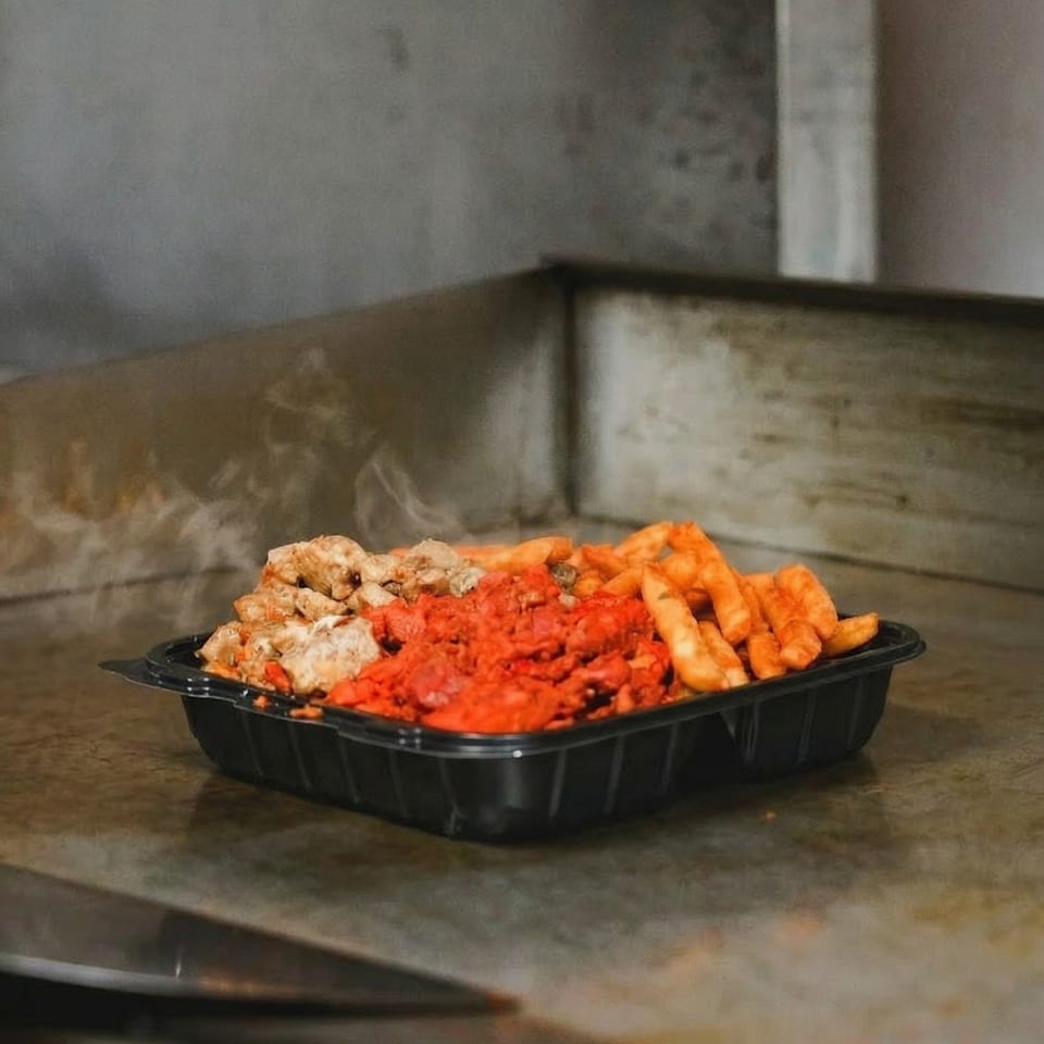 Steaming hot chicken, red spiced meat, and fries in a black takeout container on a griddle.