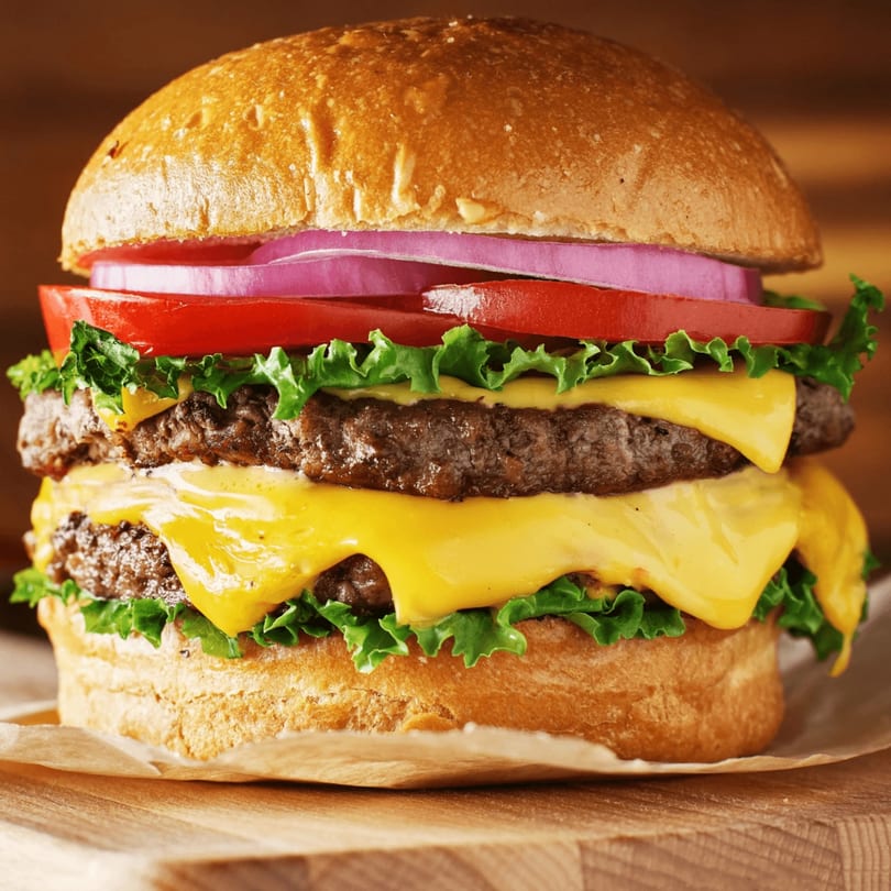A double cheeseburger with lettuce, tomato, and red onion on a bun, on a wooden surface.